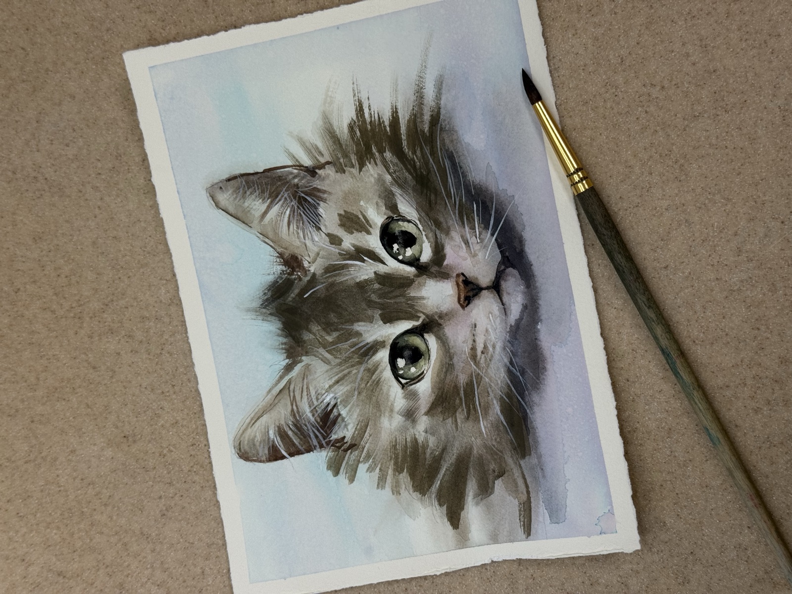 Watercolor portrait of a wide-eyed tabby kitten on paper beside a paintbrush
